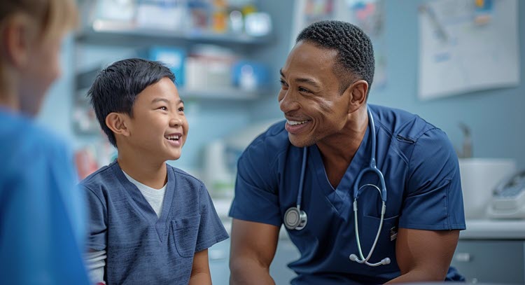 An African American doctor smiles at an Asian child patient, with both wearing blue.