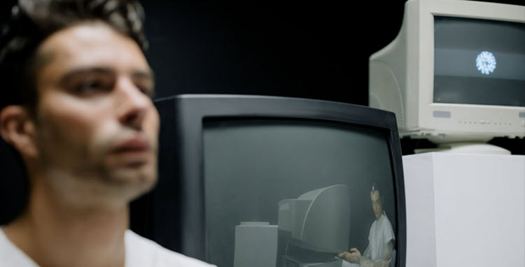 Individual positioned near computer screens displaying monitored images, representing concerns about ambient documentation, AI-assisted recording, patient privacy, and how awareness of observation may influence communication.