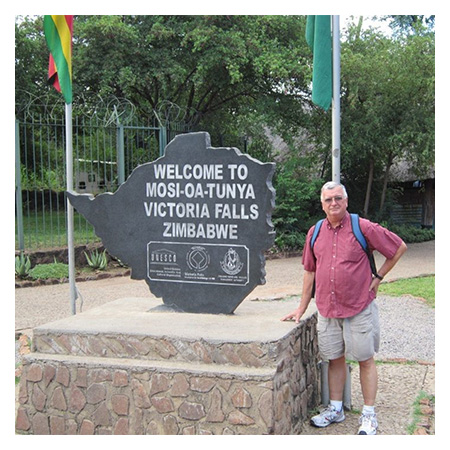 Jim Pollard standing next to a sign saying Welcome to Mosi-Oa-Tunya, Victoria Falls, Zimbabwe with a Zimbabwean flag and trees in the background