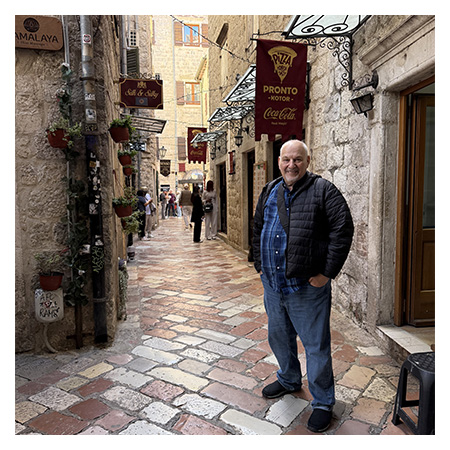 Gary Baum on a narrow cobblestone street lined with stone buildings and hanging restaurant signs in Kotor.