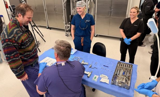 Orthopedic faculty and trainees stand around a surgical workstation displaying orthopedic instruments and implants during an educational cadaver lab session.