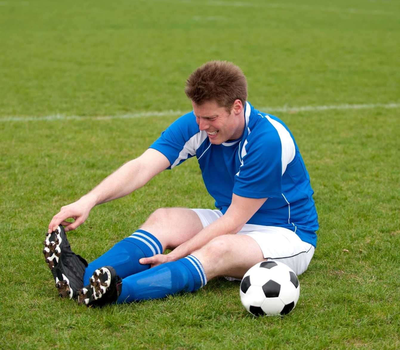 Soccer player sitting on the field holding his injured ankle after a rolled ankle during a match.