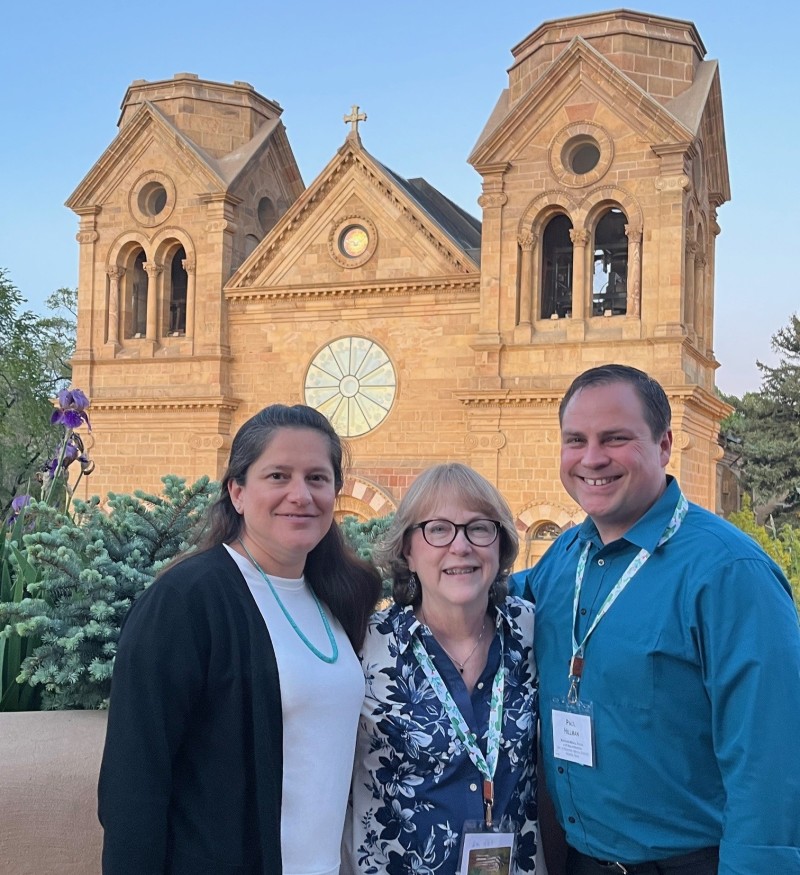 Three people smile in front of a brick cathedral