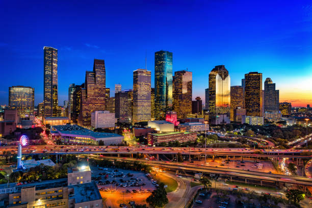 An elevated aerial view of Downtown Houston skyline at dusk with sunset reflections, and with the Gulf Freeway (I-45) and streets in the foreground.