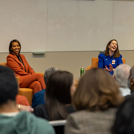 Woman with brown hair in orange pant suit sits to the left of woman with brown hair in blue dress at the front of an auditorium.