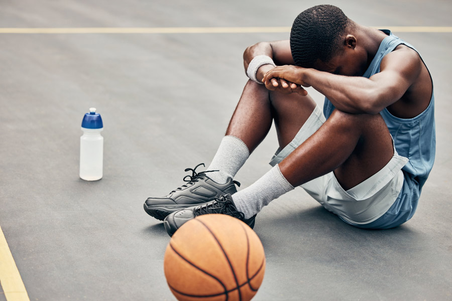 A basketball player sits on an outdoor court with his head resting on his arms, appearing fatigued or disappointed, with a basketball and water bottle nearby.