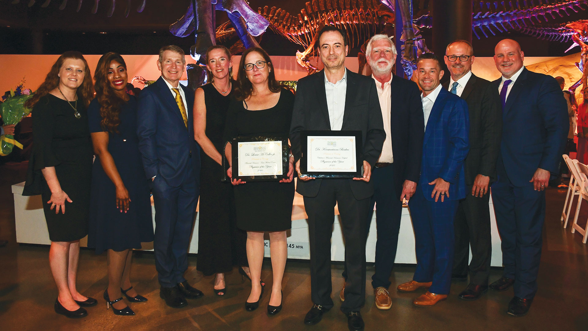 A group of nine professionally dressed adults stand together at an indoor event, smiling for a photo beneath large dinosaur skeleton exhibits. Two individuals in the center hold framed “Physician of the Year” awards, while others stand close on either side in suits and formal attire.