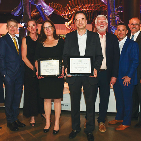 A group of nine professionally dressed adults stand together at an indoor event, smiling for a photo beneath large dinosaur skeleton exhibits. Two individuals in the center hold framed “Physician of the Year” awards, while others stand close on either side in suits and formal attire.
