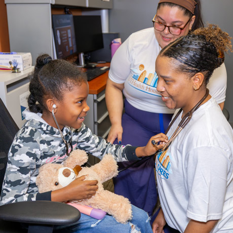 A young girl holding a teddy bear learns how to use a stethoscope at a no-cost health care screening.