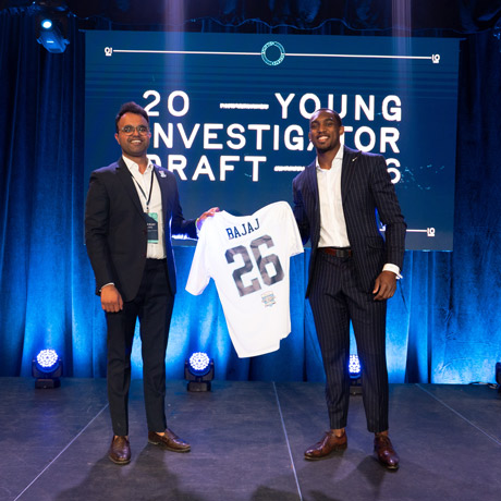 Dr. Sunanjay Bajaj receives a jersey that reads Bajaj 26 from another man dressed in a suit as the 2026 Young Investigator Draft.