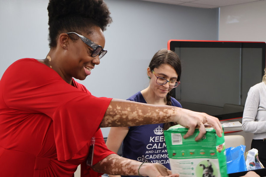 Volunteers pour pet food from a large bag into smaller containers while working together at an indoor packing station.