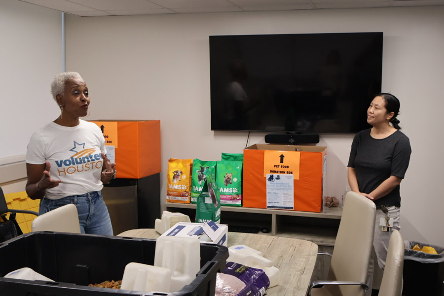 A volunteer in a “Volunteer Houston” shirt speaks to a small group, with boxes of pet food and donation bins visible in the room.