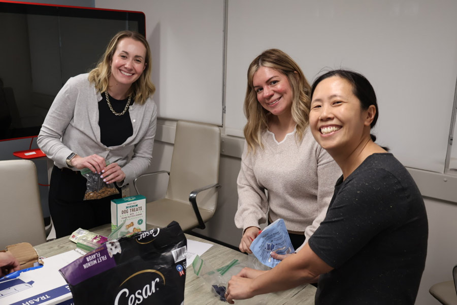 Three volunteers smile at the camera while filling small bags with dog treats during an AniMeals packing session.