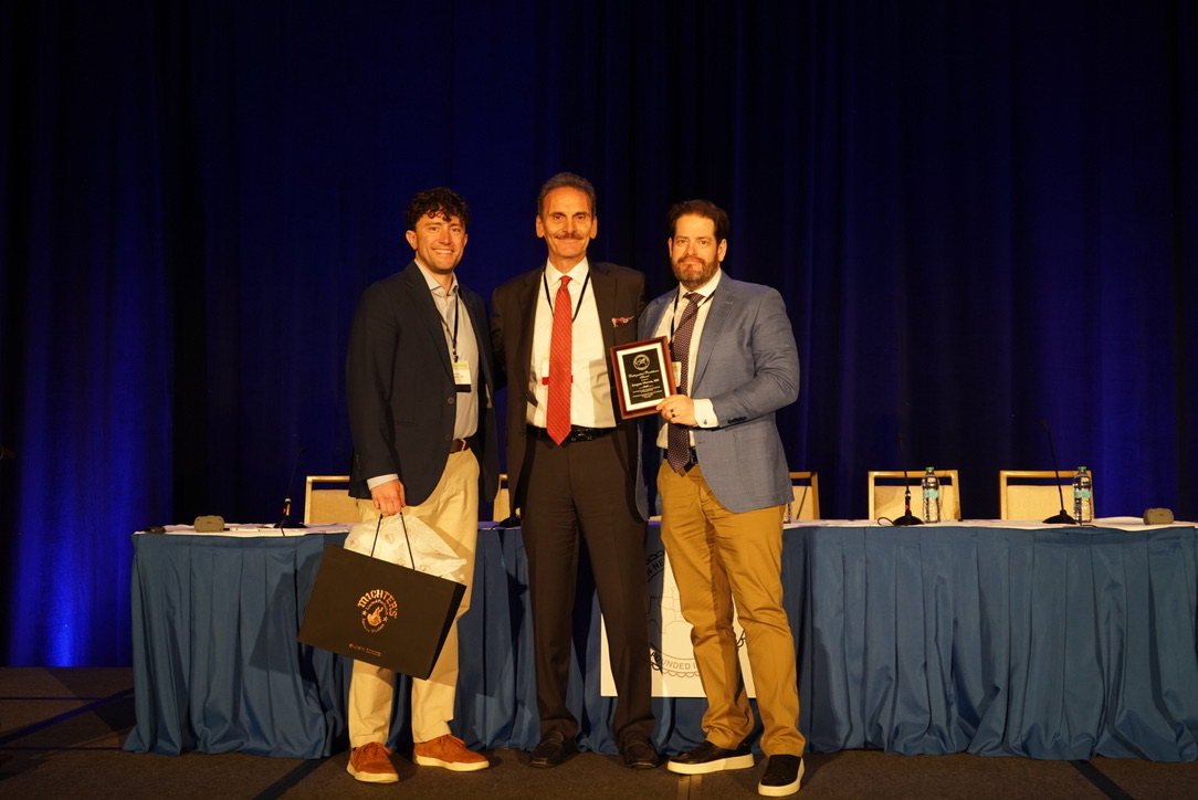 Dr. Jacques Morcos stands on a stage between two colleagues as he receives an award during a Southern Neurosurgical Society event. Morcos, dressed in a dark suit with a red tie, holds a plaque while the two men beside him—one holding a gift bag—pose for a photo. A long table with microphones and water bottles sits behind them against a blue curtain backdrop.