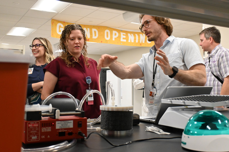 Faculty and staff gather around lab equipment during a demonstration, with one man gesturing as he explains a device to colleagues beneath a “Grand Opening” banner.