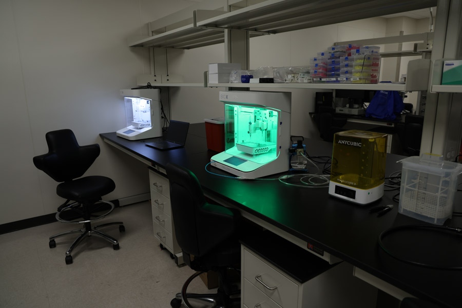 Laboratory workspace with multiple benchtop 3D printers in operation, including one illuminated in green, surrounded by research supplies, tubing, and storage containers.