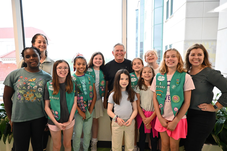 Girl Scouts from Troop 147992 pose for a group photo with adult leaders and a hospital staff member in a bright hospital atrium.