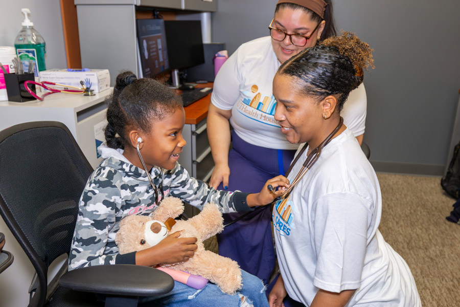 A young girl holding a teddy bear learns how to use a stethoscope at a no-cost health care screening.