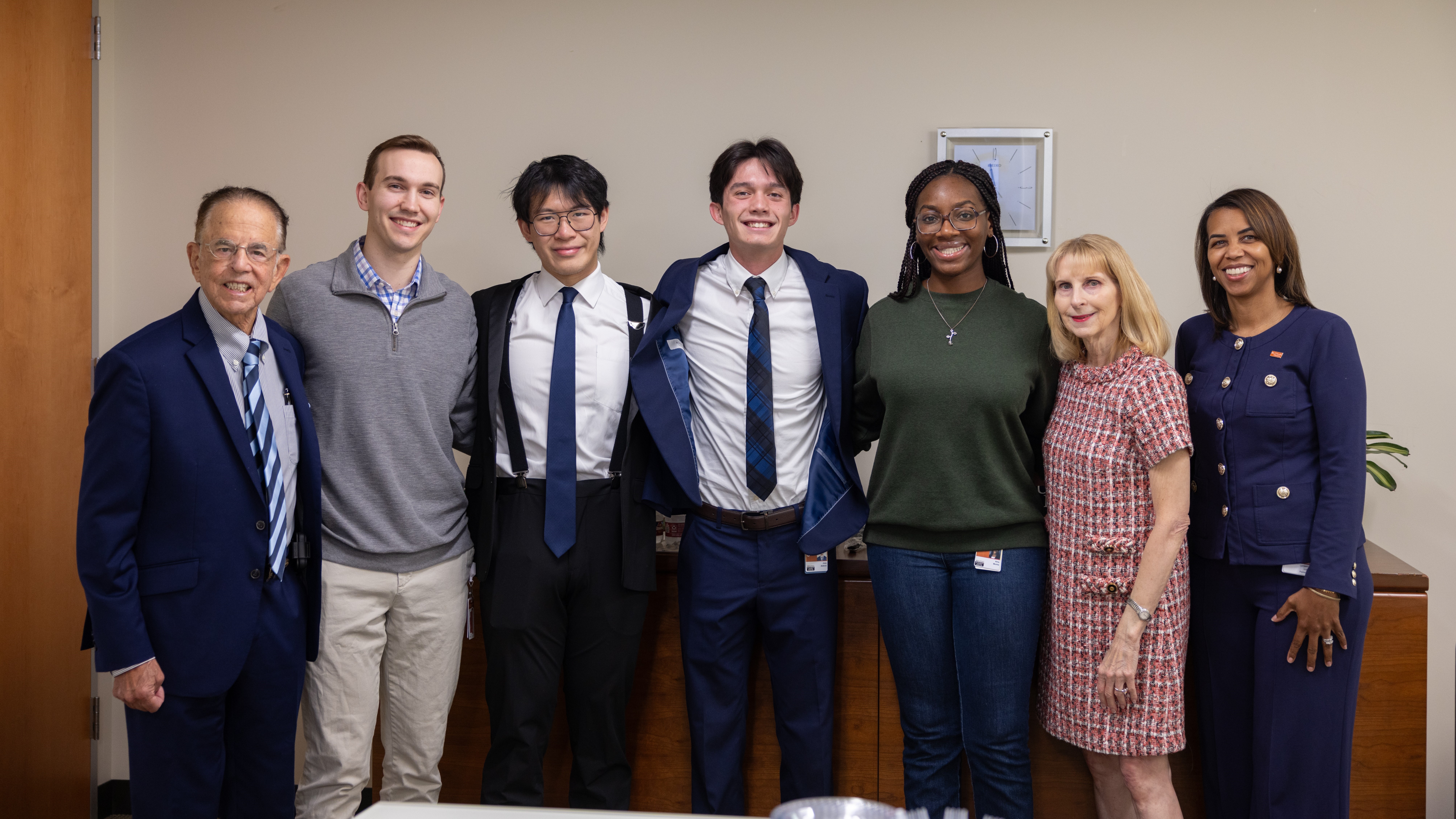 A group of seven people dressed in business clothing gather for a group photo to celebrate the recipient of a scholarship.