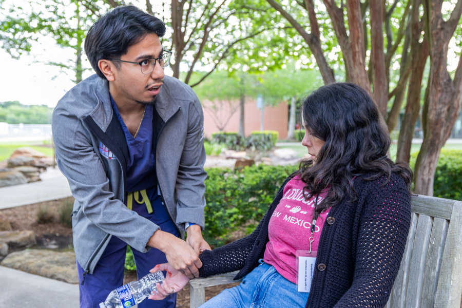 A McGovern Medical School student in medical scrubs and a jacket kneels beside a woman seated on a bench outdoors, checking her wrist as she holds a water bottle during a mass casualty simulation. The man is checking the woman's pulse.