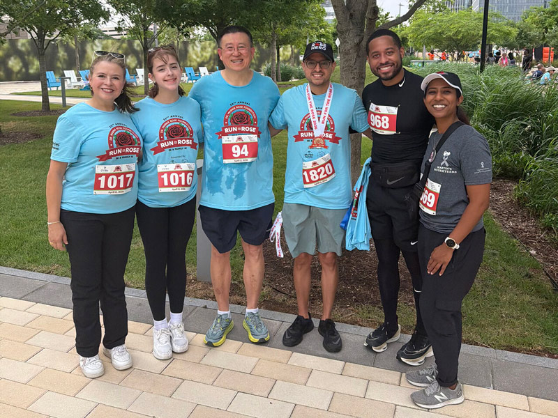 Six participants stand together outdoors after the Run for the Rose event, smiling and posing in a park-like setting with trees and a paved walkway. Most wear matching light blue “Run for the Rose” T-shirts and race bibs, while one participant in black athletic wear and another in a gray T-shirt also display bib numbers. The group appears relaxed and celebratory, with greenery and buildings visible in the background.