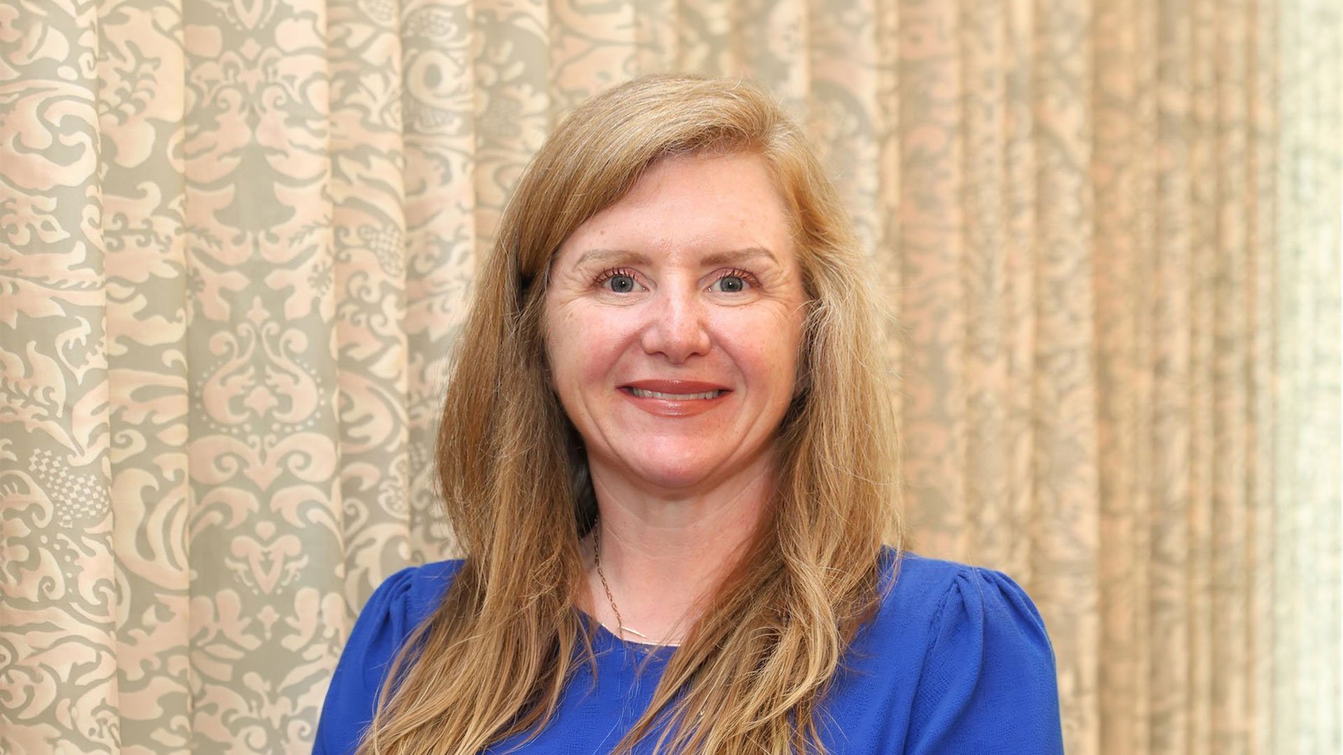Portrait of Sarah Wall with long blonde hair wearing a royal blue blouse, smiling in front of a patterned beige curtain backdrop.