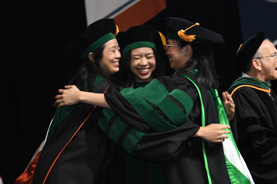 Graduates in academic regalia embrace and smile on stage during a commencement ceremony as another faculty member applauds nearby.