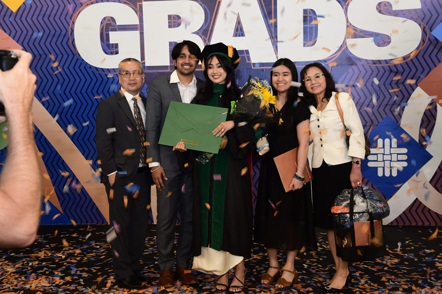 A graduate poses with family members in front of a celebration backdrop following commencement ceremonies at NRG Arena.