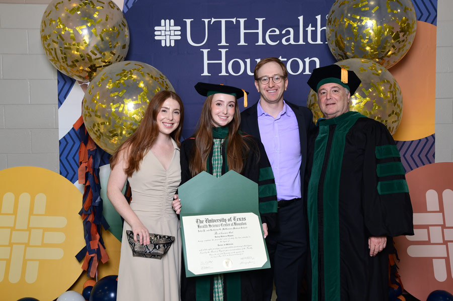 A graduate poses with family members and a diploma in front of a UTHealth Houston celebration backdrop.