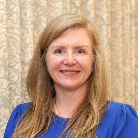 Portrait of Sarah Wall with long blonde hair wearing a royal blue blouse, smiling in front of a patterned beige curtain backdrop.