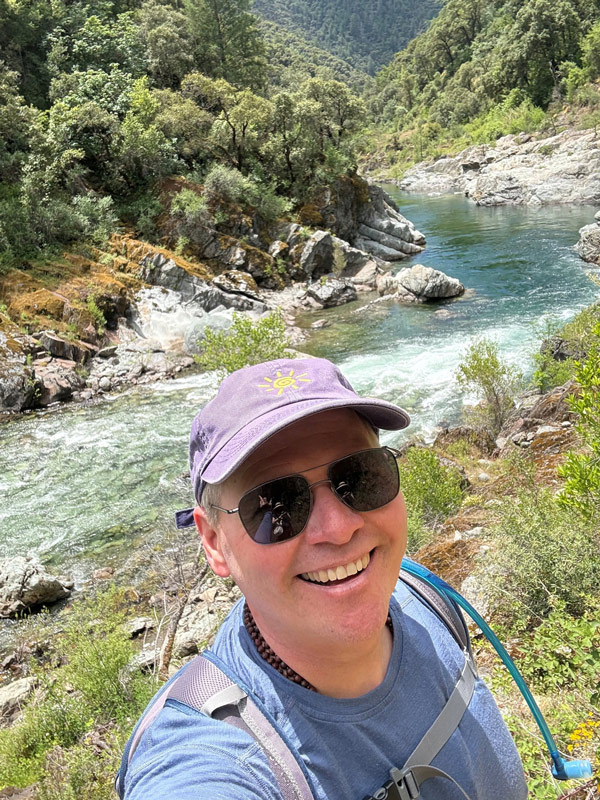 Dr. Ed Fink, wearing a blue shirt and a purple hat stands in front of a river and a mountain.