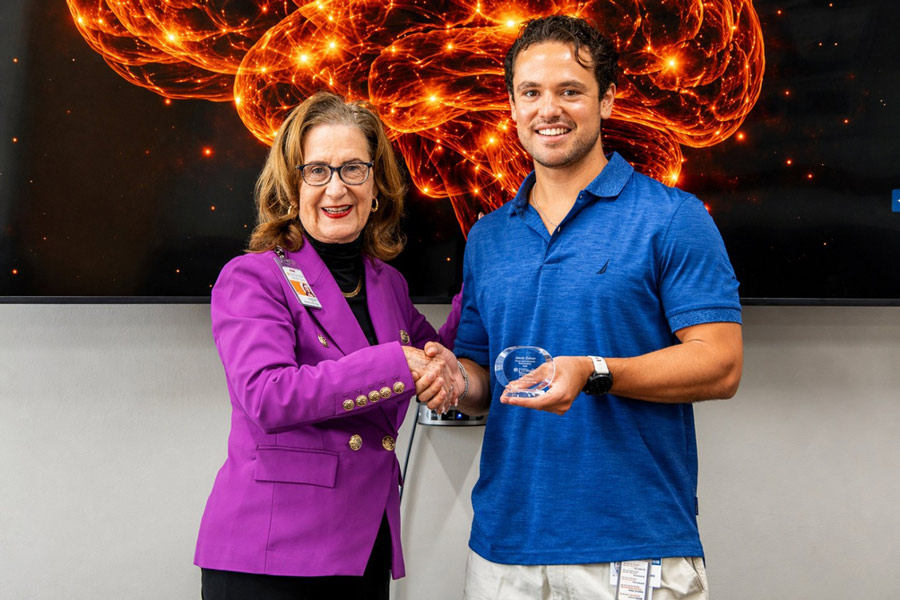 Jacob Galan, dressed in a blue polo receives an award from Dr. Claire Hulsebosch, dressed in a purple blazer with black undershirt, in front of a 3D image of neurons of a brain