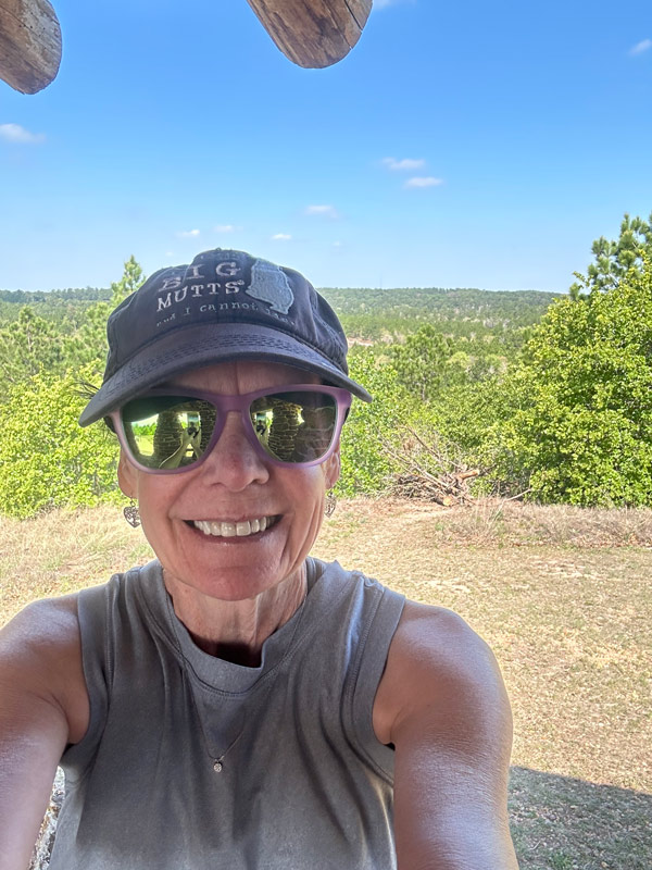 Jill Herrin wearing a gray shirt and a blue hat poses in front of the landscape of Bastrop State Park.