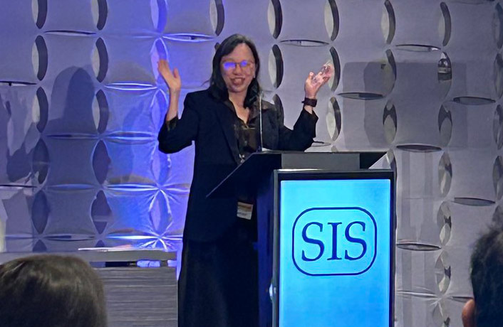 Dr. Lillian Kaw speaks at a podium during the SIS annual conference presentation, gesturing with both hands while addressing the audience.
