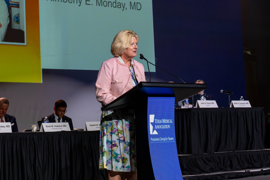 Kimberly Monday, MD, speaks at a podium during the Texas Medical Association annual conference while seated physician leaders look on from the stage behind her. A large presentation screen is visible in the background, and the Texas Medical Association logo is displayed on the podium.