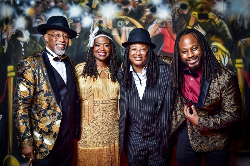 The four members of the Kincade family dressed in 1920s era clothing, posting in front of a paparazzi type backdrop