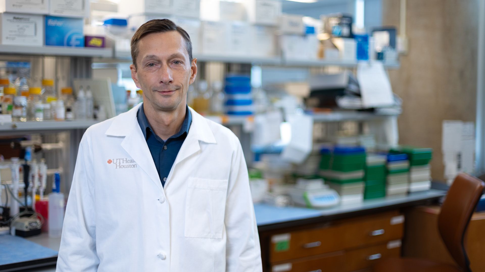 Researcher in a UTHealth Houston lab wearing a white lab coat stands in front of laboratory benches filled with scientific equipment, bottles, and storage containers.