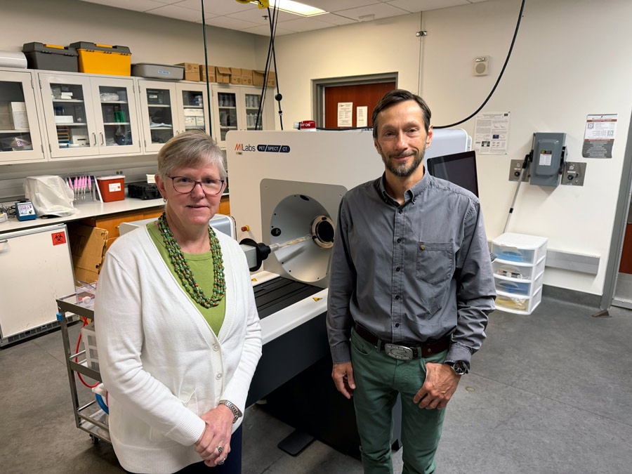 Eva Sevick and Mikhail Kolonin stand in a laboratory in front of imaging equipment and research workstations. Sevick wears a white cardigan and green necklace, while Kolonin wears a gray button-down shirt and green pants. Cabinets filled with lab supplies line the background.
