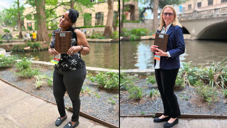 Two women stand outdoors beside the San Antonio River Walk holding award plaques during the Laboratory Management Association Annual Meeting. The woman on the left wears a black floral top and black pants, while the woman on the right wears a navy blazer and black pants. Trees, water, and a stone bridge are visible in the background.