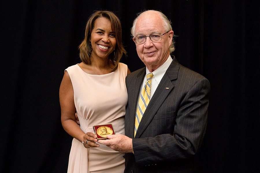 Two individuals pose with a gold medallion in a red case, one presenting the award to the other in front of a black curtain.