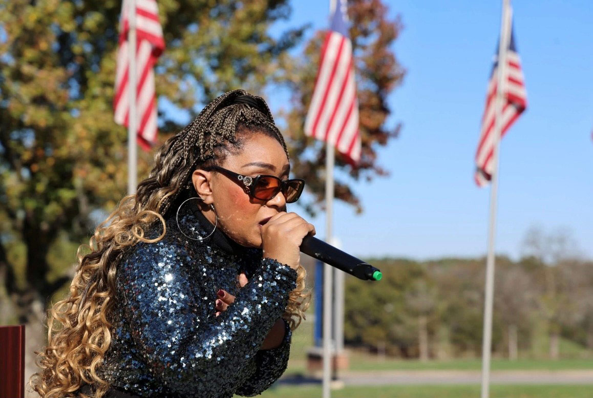 Performer wearing a sequined jacket and sunglasses sings into a microphone outdoors with American flags displayed in the background.