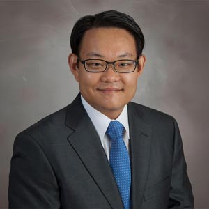 Dr. H. Alex Choi poses for a headshot in a gray suite, blue tie, and white shirt in front of a light gray backgroudn.