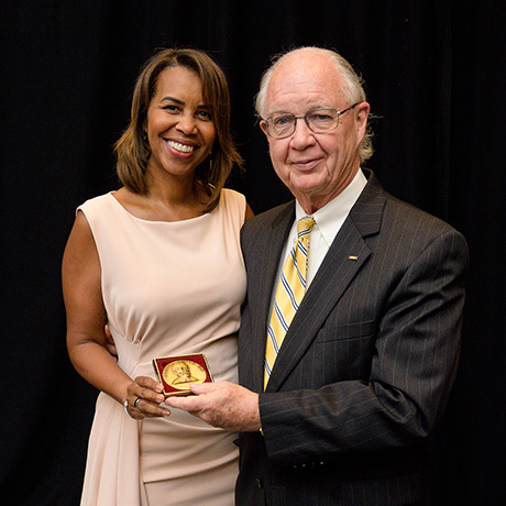 Two individuals pose with a gold medallion in a red case, one presenting the award to the other in front of a black curtain.