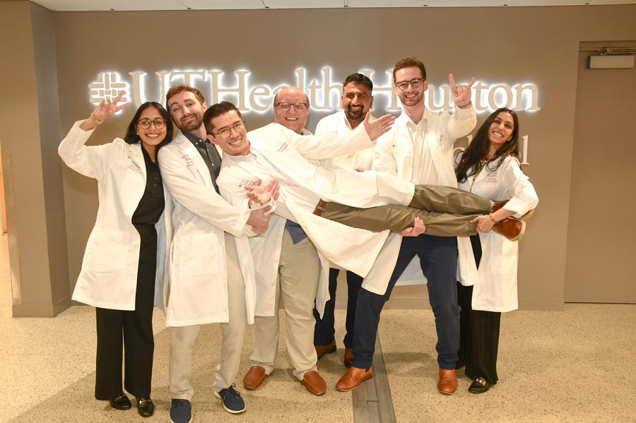 Seven McGovern Medical School internal medicine interns in white coats pose for a playful group photo after headshot day, holding one intern horizontally in front of a UTHealth Houston sign before officially arriving on campus.