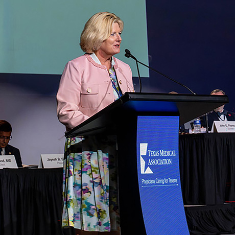 A woman in a pink jacket speaks at a podium during a Texas Medical Association meeting, while seated panelists listen from a stage