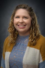 Professional headshot of Dr. Autumn Marsden smiling at the camera against a dark studio background. She is wearing a blue lace blouse with a color-block cardigan and has shoulder-length curly hair.