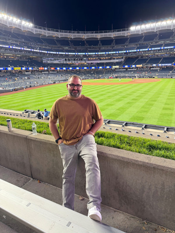 A person in a brown shirt and grey jeans standing in the front row of a baseball stadium overlooking the field at night, posing near the outfield seating area with stadium lights illuminating the ballpark in the background.