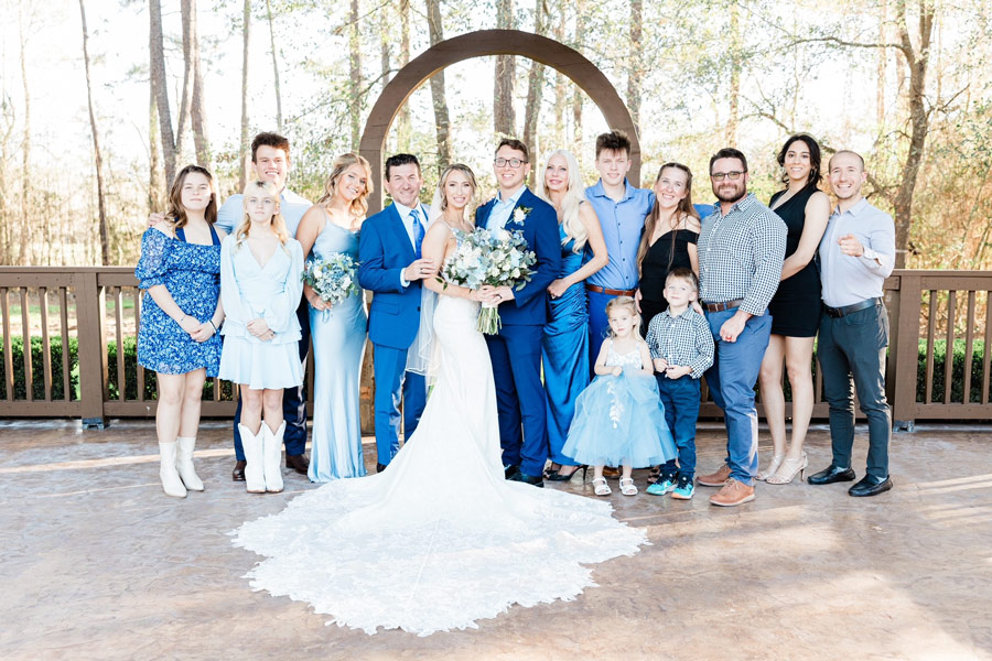 Large family group poses outdoors for a wedding portrait with the bride and groom centered beneath a wooden arch.