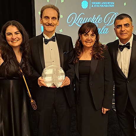 Four formally dressed people pose together at a gala event, with the man in the center holding a clear award. A stage backdrop behind them