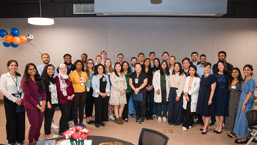 Group portrait of the 2026 graduating cohort of the Physician Educator Certificate Program standing together indoors during a recognition event. Residents, fellows, faculty, and program leaders pose in several rows, with participants wearing a mix of professional attire, white coats, and clinical scrubs beneath blue and orange decorations celebrating the occasion.